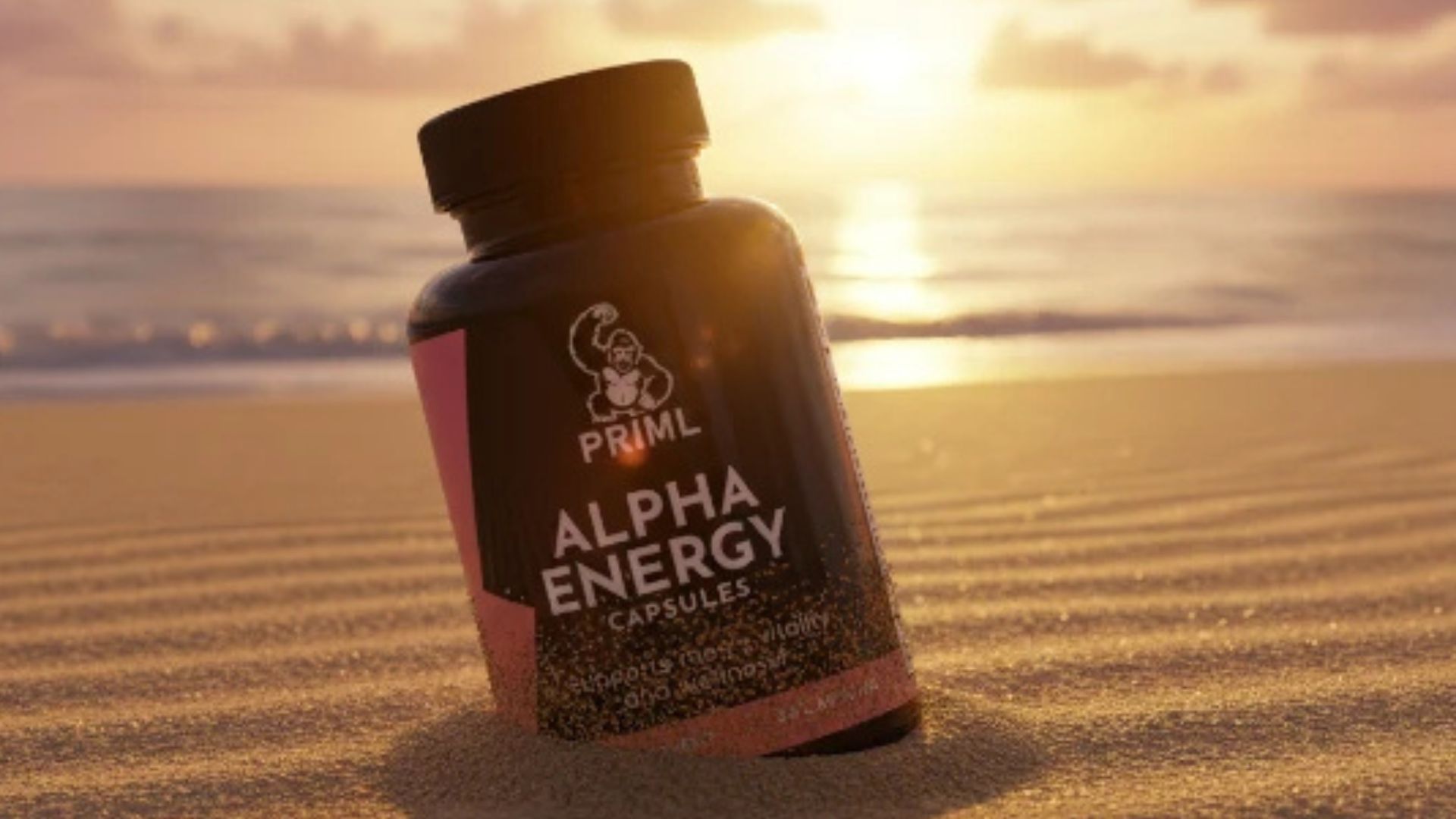 A bottle of PRML Alpha Energy capsules stands upright in the sand on a beach at sunset, with the ocean and sun in the background.